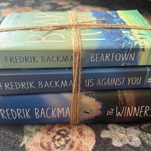 May include: A stack of three books by Fredrik Backman titled Beartown, Us Against You, and The Winners, tied together with a piece of brown twine. The books feature blue and green covers with white text and are stacked vertically on top of each other.