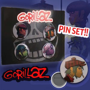 Puede incluir: Un set de pines de Gorillaz con cuatro chapas circulares sobre una tarjeta negra con un gráfico de calavera. Los pines muestran personajes ilustrados de la banda. El texto GORILLAZ y PIN SET!! aparece en el embalaje.