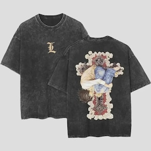 May include: A charcoal gray acid-wash t-shirt featuring a small gold letter L on the front chest. The back displays a large graphic of a person with dark hair curled up inside a decorative cross adorned with red and white floral patterns.