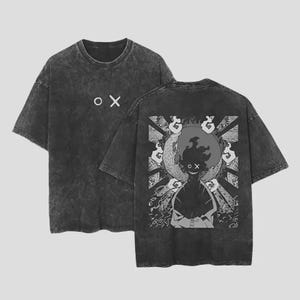 May include: A charcoal grey acid-wash t-shirt featuring a small white circle and cross symbol on the front chest. The back displays a large graphic of a silhouetted figure with a wide grin, surrounded by a moon, sunburst rays, and decorative patterns.