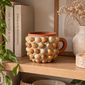 Op de afbeelding: Een terracotta mok met een afgerond handvat, waarvan het buitenoppervlak dicht is bedekt met kleine, getextureerde schelpen in tinten bruin, beige en crème. De rand en het handvat zijn van glad, onbewerkt terracotta in een warme roodbruine kleur.