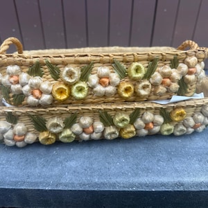 May include: A set of two rectangular woven straw baskets featuring rope handles and decorative floral accents. The baskets are adorned with light-colored straw flowers, green leaves, and small orange centers, creating a textured, natural appearance.