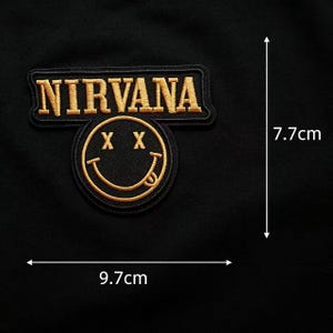 May include: A black embroidered patch featuring the word NIRVANA in gold thread above a gold smiley face with X eyes and a tongue sticking out. The patch measures 9.7 cm in width and 7.7 cm in height as indicated by the visible measurement lines.
