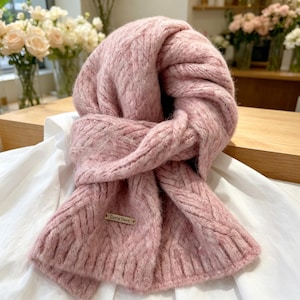 May include: A soft, chunky knit scarf in a dusty pink hue featuring a textured herringbone pattern throughout. The scarf has a ribbed hem and a small, rectangular metallic tag attached to the front with the engraved text Daria Dore.