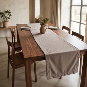 May include: A long rectangular beige fabric table runner centered on a dark brown wooden dining table. The table is surrounded by four matching wooden chairs and features a ceramic pitcher with dried flowers and two place settings.