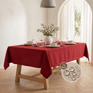 May include: A solid deep red rectangular tablecloth draped over a wooden table. The table is set with white plates, clear glasses, and a central vase with green branches. An inset circle shows water droplets beading on the fabric surface.