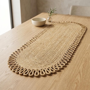 May include: An oval-shaped table runner crafted from natural tan woven fibres. The runner features a dense, coiled centre section surrounded by a decorative border of repeating, looped fibre strands that create a scalloped edge effect.
