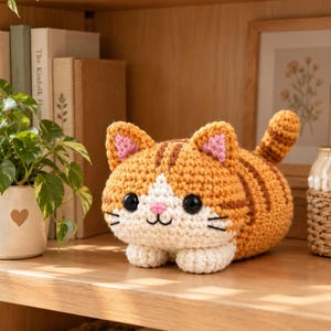 May include: A crocheted orange and white tabby cat plush toy with dark brown stripes, large black plastic eyes, a small pink stitched nose, and pink inner ears. The cat is in a lying position with its tail raised and white paws tucked underneath.