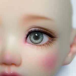 May include: Close-up of a doll's face with green eyes. The doll has pink blush on her cheeks and a light pink lip. The text "12/5 mm (small iris) www.nomyens.com" is visible below the doll's face.
