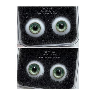 May include: Two pairs of doll eyes with green irises and white sclera, presented in a black foam-lined case. The text "18/7 mm (Small iris) www.nomyens.com" is visible. These eyes are designed for dolls or similar crafts.