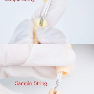 May include: Close-up of a white string being threaded through a small hole in a doll's arm. The string is attached to a metal hook.