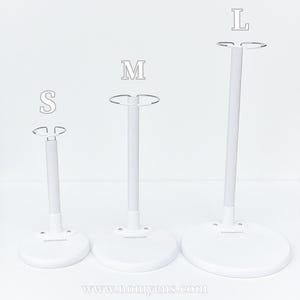 May include: Three white doll stands in sizes S, M, and L. Each stand has a round base, a vertical pole, and a wire loop at the top. The stands are designed to hold dolls and are displayed against a white background.