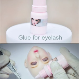 May include: A small pink bottle of eyelash glue with a doll's face on the label. The bottle is on a white surface with the text "Glue for eyelash" written below it.