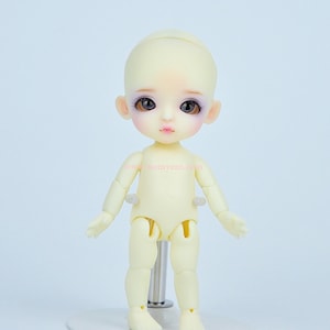 Puede incluir: Una pequeña muñeca articulada amarilla con ojos marrones y sombra de ojos morada. La muñeca está de pie sobre un soporte blanco.