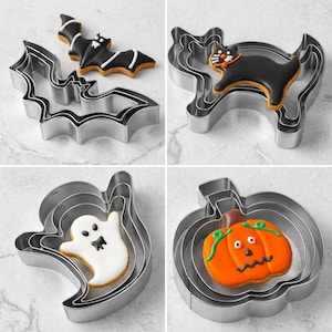 Puede incluir: Un conjunto de cuatro colecciones de cortadores de galletas de metal con formas de Halloween, incluyendo un murciélago, un gato, un fantasma y una calabaza. Cada set contiene cortadores anidados con una galleta decorada encima.