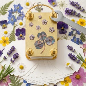 May include: A rectangular gold-toned flower press featuring a central four-leaf clover illustration and small floral accents. The device is secured with four corner bolts and a top ring, with a silver wrench positioned on one bolt.