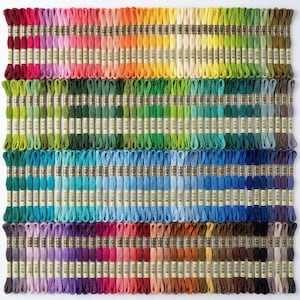 May include: A large collection of embroidery floss skeins arranged in a grid pattern. The skeins display a full spectrum of colours including reds, oranges, yellows, greens, blues, purples, browns, greys, and blacks, each with a paper label.