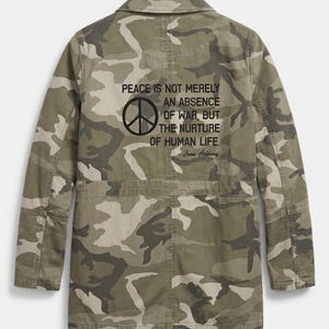 May include: A camouflage print jacket featuring a collar, long sleeves, and a back graphic with a peace symbol and the text: PEACE IS NOT MERELY AN ABSENCE OF WAR, BUT THE NURTURE OF HUMAN LIFE. Jane Addams. The jacket has a horizontal seam at the waist.