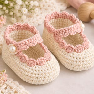 May include: A pair of cream-colored crochet baby shoes featuring a scalloped pink trim along the ankle strap and a single white faux pearl button on the side of each shoe. The shoes are crafted with a dense stitch pattern and a rounded toe design.