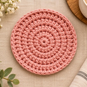 May include: A circular, dusty pink crochet coaster featuring a dense, concentric stitch pattern. The coaster is crafted from thick yarn, displaying a uniform, textured surface with a central starting point and radiating rows of stitches.