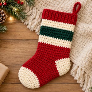 Pode incluir: Uma meia de Natal de crochê feita à mão com um punho vermelho, uma faixa creme, uma faixa verde escura e uma segunda faixa creme. O corpo principal é vermelho com calcanhar e ponta creme, e uma alça vermelha para pendurar.