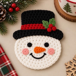 May include: A round, white crocheted snowman coaster featuring a black top hat with a red band, green holly leaves, and three red berries. The snowman has black circular eyes, a curved black mouth, an orange carrot nose, and pink cheeks.