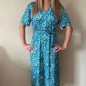 May include: A vintage-style midi dress in a blue floral pattern. The dress features short sleeves, a V-neck, and a matching belt that cinches the waist. The fabric has a light, airy appearance, perfect for warmer weather.