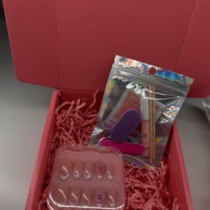 Könnte beinhalten: Eine rosa Geschenkbox mit einem durchsichtigen Kunststoffetui, das zehn künstliche Fingernägel mit weißen Spitzen und Blumenverzierungen enthält. Zudem liegt ein holografischer Beutel mit einem lila Buffer, einer rosa Feile, Nagelkleber und zwei Stäbchen bei.
