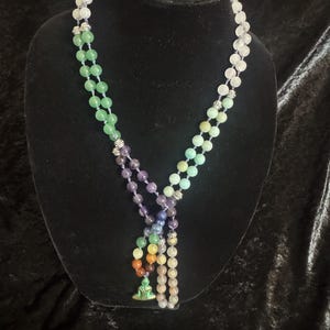 May include: A long beaded necklace featuring a variety of smooth, round, multi-colored gemstone beads arranged in sections of green, purple, white, and pale blue. The necklace includes silver-toned metal spacers and a small green Buddha pendant.