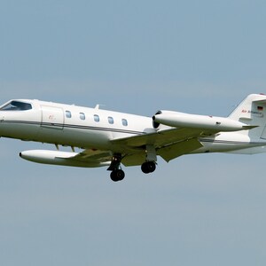 May include: A white private jet in flight with landing gear extended. The aircraft features a sleek fuselage, a T-tail configuration, and two rear-mounted engines. The text Air Alliance is visible on the tail section near a small German flag.