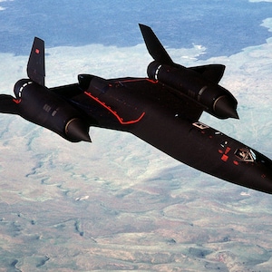 May include: A sleek black supersonic reconnaissance aircraft flying at high altitude. The jet features a long, pointed nose, two large engine nacelles mounted on delta wings, and two vertical stabilizers. Thin red lines accent the fuselage.