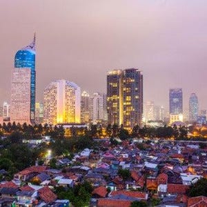 May include: A cityscape featuring a dense collection of low-rise buildings with red-tiled roofs in the foreground, contrasted against a backdrop of modern skyscrapers and high-rise towers under a soft, hazy, purple-tinted twilight sky.