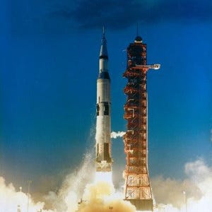 May include: A tall white and black Saturn V rocket launching into a deep blue sky, emitting bright orange flames and thick white smoke at the base. Beside the rocket stands a tall, red, metal launch umbilical tower structure.