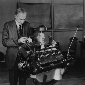 May include: A black and white photograph showing a person in a suit and tie working on a large internal combustion engine. The engine features a prominent air cleaner, a carburetor, spark plugs, and a fan belt assembly mounted on a stand.