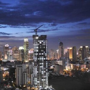May include: A cityscape at twilight featuring a dense cluster of illuminated skyscrapers and high-rise buildings. A tall building under construction with a crane on top stands prominently in the centre against a dark blue cloudy sky.