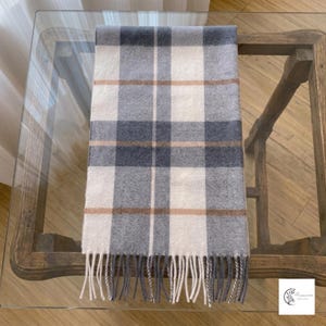 Puede incluir: Una bufanda rectangular con un patrón de cuadros en tonos blanco, gris oscuro y gris claro, acentuado por finas rayas horizontales y verticales de color beige. La bufanda tiene una apariencia suave y texturizada con flecos largos.