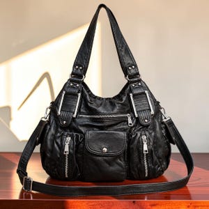 Puede incluir: Un bolso de hombro negro con acabado texturizado, con dos asas superiores y una correa cruzada desmontable. La parte frontal incluye un bolsillo central con solapa y broche plateado, dos bolsillos laterales verticales y un compartimento con cremallera.