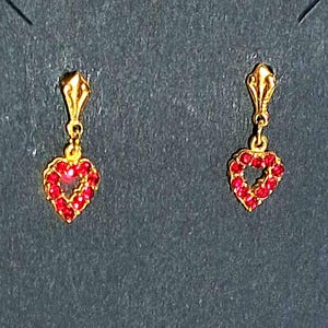May include: A pair of gold-toned drop earrings featuring a small, dangling heart-shaped charm. Each heart is outlined with tiny, vibrant red stones. The earrings are attached to a simple gold-toned post that connects to the heart charm via a small loop.