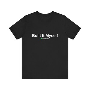 以下が含まれることがあります： 胸の中央に白い文字で「Built It Myself」とプリントされた黒の半袖Tシャツ。文字の下には小さな緑色の円と白い文字で「CABLECRED」が配置されています。クルーネックで標準的なフィット感のTシャツです。
