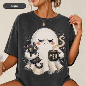 Könnte beinhalten: Ein pfefferfarbenes T-Shirt mit einer Grafik eines mürrischen weißen Geistes, der eine schwarze Katze und eine Tasse mit der Aufschrift MEH hält. Der Geist hat einen finsteren Ausdruck und rote Wangen, umgeben von kleinen Sternen und Monden.