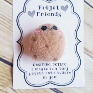May include: A plush, brown potato-shaped fidget toy with black eyes and pink cheeks, presented on a white card. The card reads "Fidget Friends" and "Positive Potato: I maybe be a tiny potato but I believe in you!"