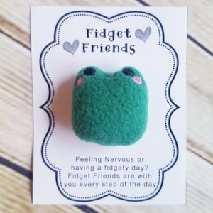 May include: A green, textured frog-shaped fidget toy with pink cheeks, presented on a white card. The card features the text "Fidget Friends" and a message about managing anxiety. The background is a light wood grain.