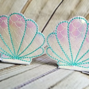 May include: Two iridescent white seashell hair clips with teal stitching. The seashells are designed with a mermaid scale pattern.