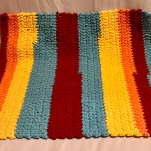 May include: A rectangular hand-knitted blanket featuring thick, chunky yarn in a repeating vertical striped pattern. The colors include vibrant red, bright yellow, warm orange, and light blue, creating a bold, multi-colored textile design.