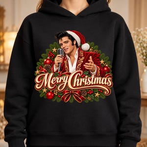 May include: A black hooded sweatshirt featuring a graphic of Elvis Presley wearing a Santa hat and a red velvet jacket, holding a microphone. The design is surrounded by a Christmas wreath with red ornaments, pinecones, and the text Merry Christmas.