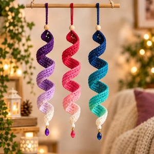 May include: Three hanging crochet spirals suspended from a horizontal wooden dowel. Each spiral features a gradient color transition from dark at the top to white at the bottom, accented with a wooden bead and a teardrop-shaped pendant at the base.