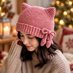 May include: A dusty pink crocheted beanie featuring two pointed cat-like ears at the top. The hat has a ribbed texture and a decorative side accent consisting of a small bow with three dangling, leaf-shaped crocheted charms attached to the brim.