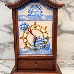 May include: A wooden mantel clock with a dark brown finish and a small drawer at the base. The clock face features a blue and white ceramic tile design depicting a ship's wheel, a sailing ship, a compass rose, and a sun with a human face.