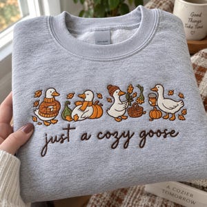 May include: A light grey crewneck sweatshirt featuring an embroidered design of four geese in autumn-themed attire and accessories, including pumpkins and leaves. Below the geese, the text "just a cozy goose" is embroidered in dark brown script.