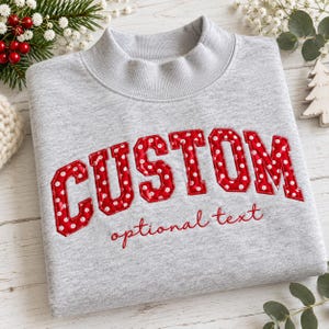 Op de afbeelding: Een lichtgrijs sweatshirt met een opstaande kraag, voorzien van het woord CUSTOM in grote, rode stoffen letters met witte stippen en gestikte randen. Onder de hoofdtekst is de tekst optional text geborduurd in een kleiner, rood cursief lettertype.